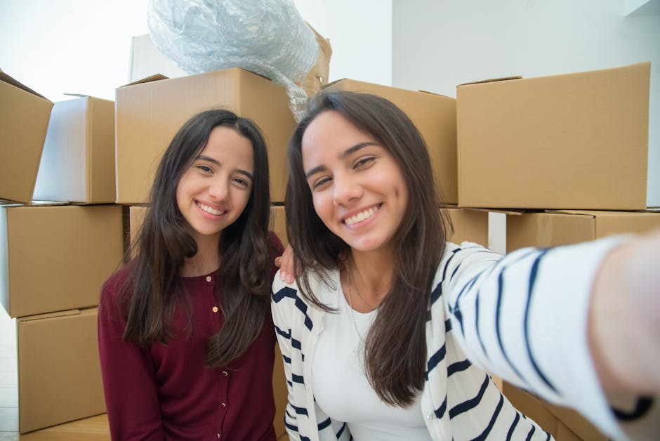 Two young women with long dark hair and smiling faces taking a selfie inside a property loaded with packed cardboard boxes of various sizes, some sealed with tape and others open, revealing packaging