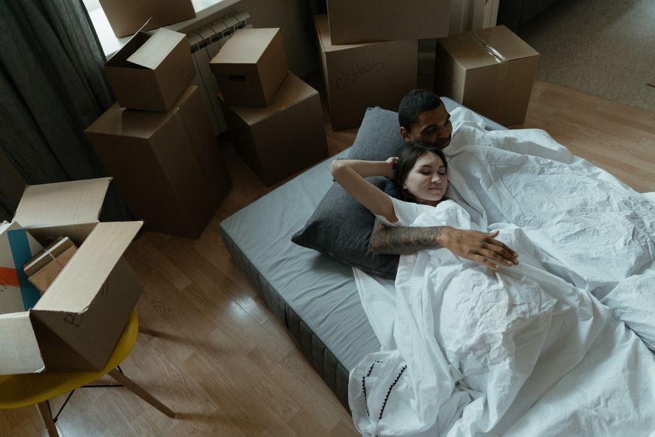 An image showing a couple lying in bed inside a room with large windows that let in natural daylight. The room contains several cardboard moving boxes placed on the windowsill and around the bed, indi